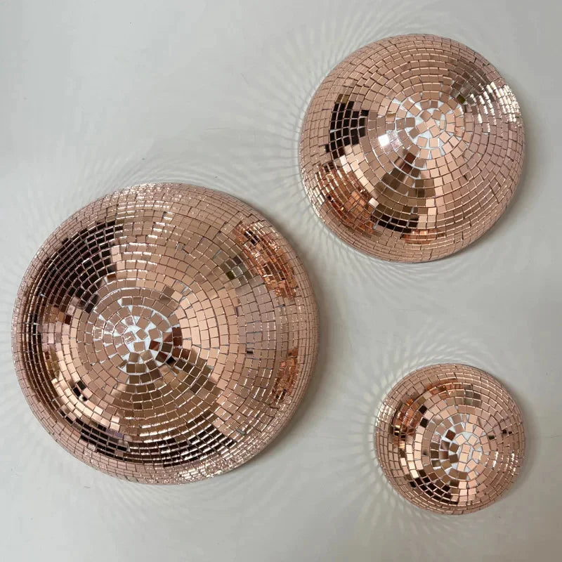 Trois miroirs ronds en or rose et boules disco reflétant la lumière sur une surface blanche.