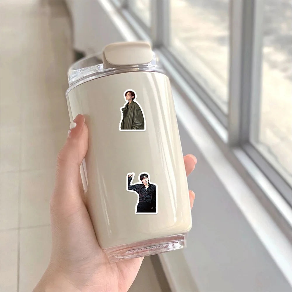 A hand holds a clear plastic bottle containing K-Pop idol stickers featuring two male idols.