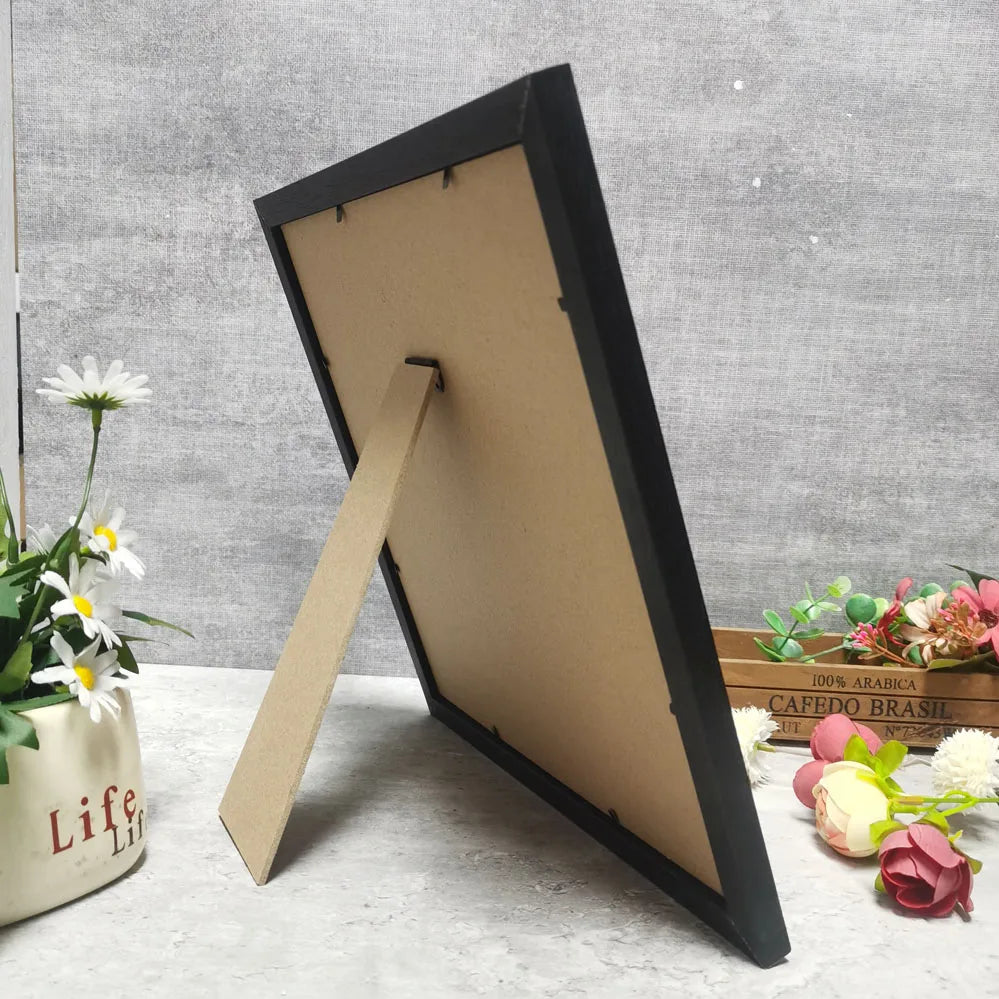 A black photo frame with a light brown backing is displayed leaning against a white surface.
