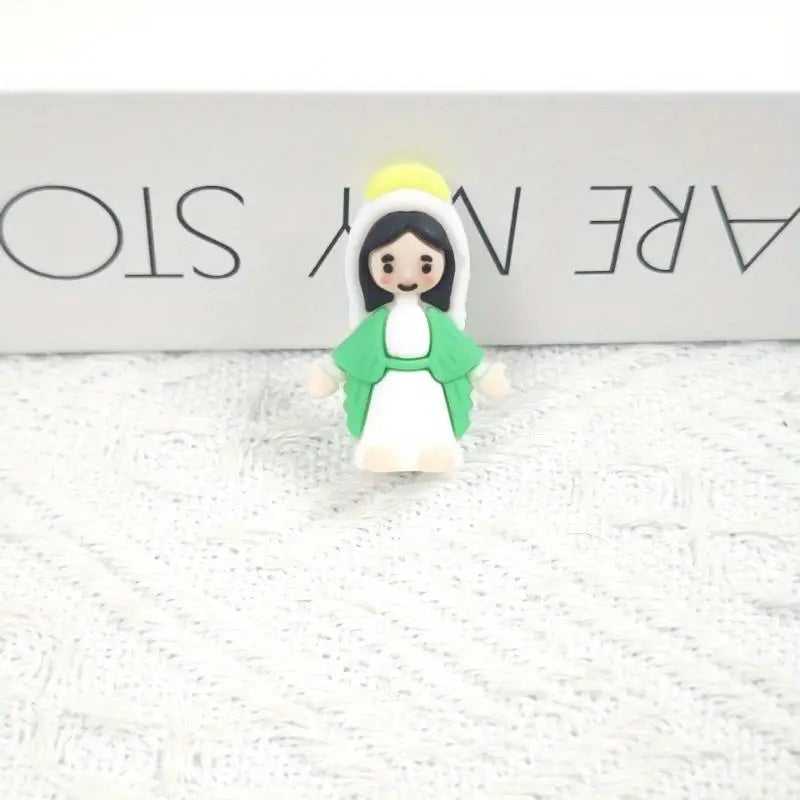 A small resin statue of the Virgin Mary with a yellow halo and green garment displayed on a white textured surface.