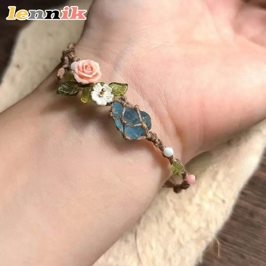 A macrame bracelet featuring a rose accent and a blue aquamarine bead worn on a hand.