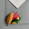 A stack of colorful food-shaped fridge magnets resembling a hamburger with a pink top and green lettuce on a gray surface.