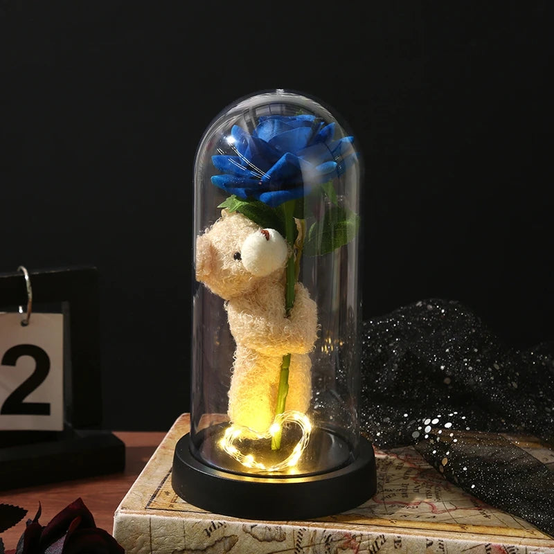 An eternal rose LED decorative flower with a blue rose inside a glass dome and a teddy bear figure.