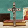 A bronze crucifix wall decor featuring a figure of Christ, an INRI plaque, and a St. Benedict medal mounted on a wooden book.
