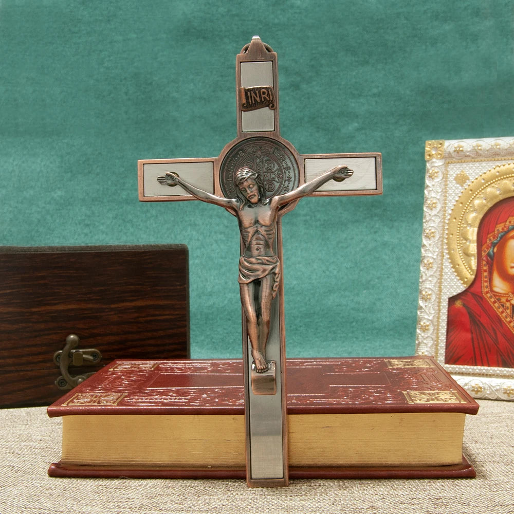 A bronze crucifix wall decor featuring a figure of Christ, an INRI plaque, and a St. Benedict medal mounted on a wooden book.