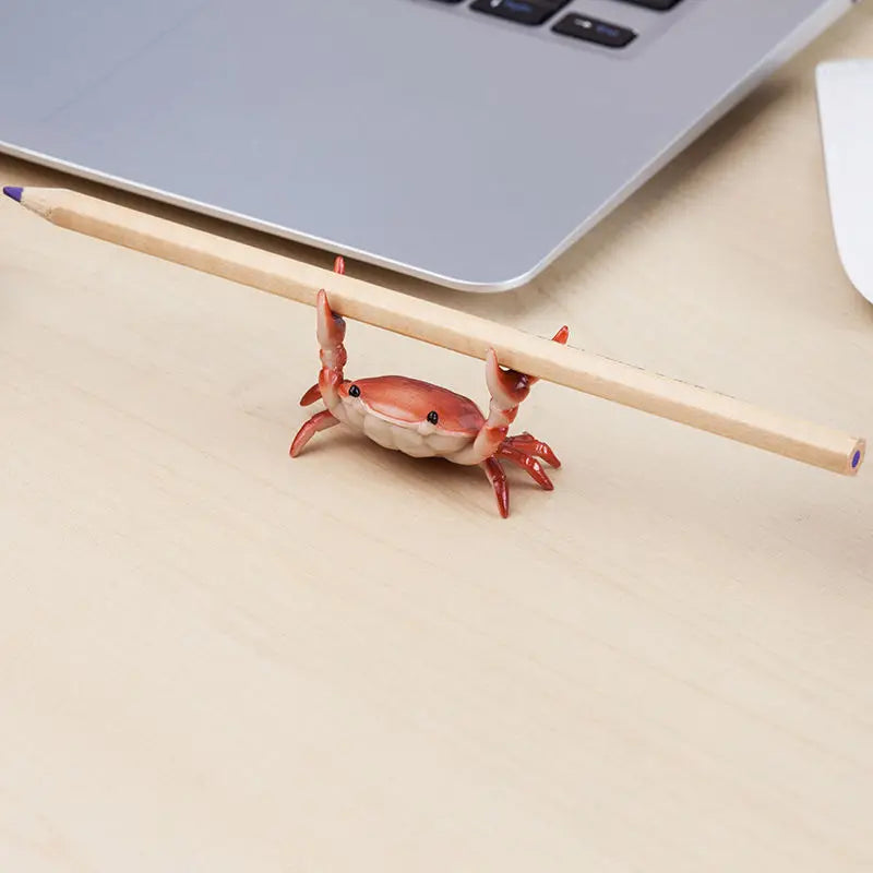 A small crab-shaped desk organizer holding a pen and phone, resting on a wooden surface next to a laptop.