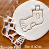 A white fondant cookie cutter shaped like a zombie foot is stamped into a white cookie on a wooden surface.