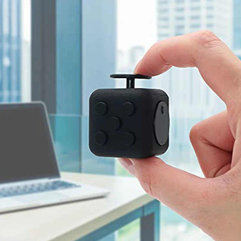 A hand holds a black fidget cube stress relief toy against a blurred modern office background.