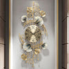 A vertical wall clock featuring a gold-toned metal design with leaf and floral elements, set against a light gray background.