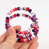 A hand holds a set of patriotic American flag beaded stretch bracelets featuring red, white, and blue beads.
