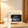 A wooden ultrasound photo frame displaying a pregnancy scan with a floral ribbon for an announcement.