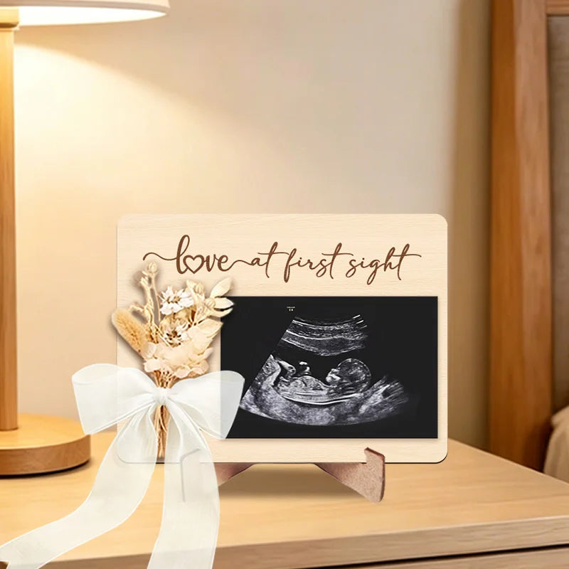 A wooden ultrasound photo frame displaying a pregnancy scan with a floral ribbon for an announcement.