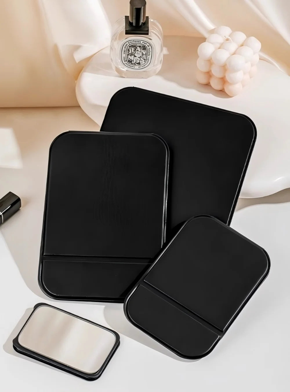 A black foldable vanity mirror with an adjustable stand and a separate compact mirror resting on a white surface.