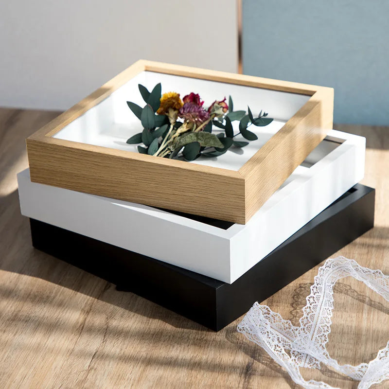 A wooden shadow box photo frame with a white interior displaying green and purple flowers, resting on a dark base.