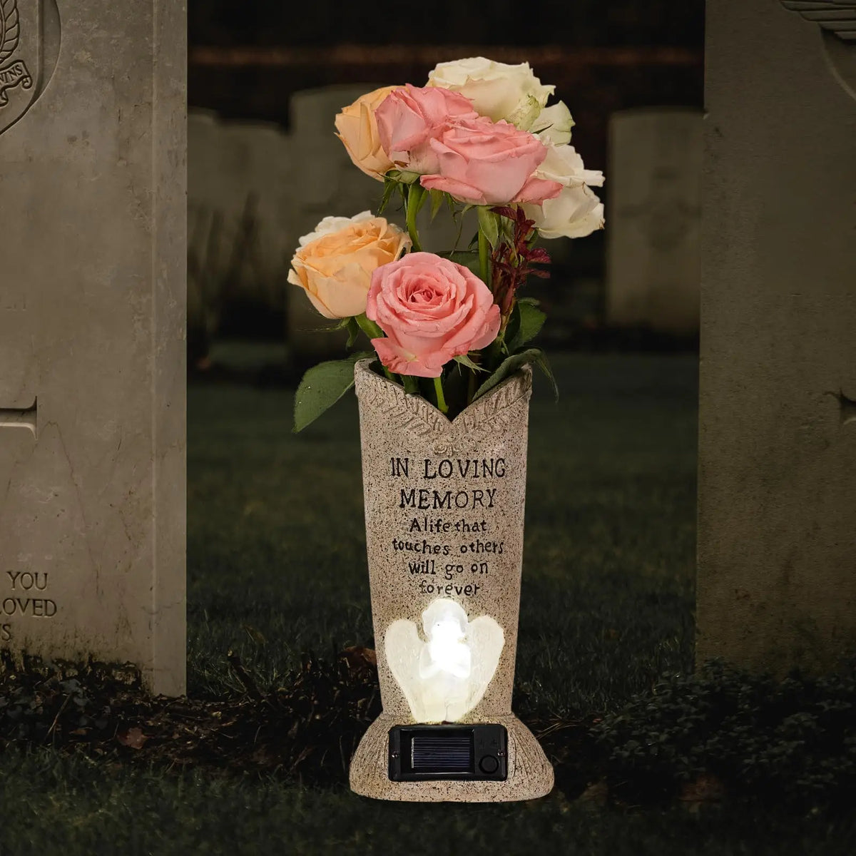 A stone vase holding pink and white roses with an LED light, placed in a cemetery setting.