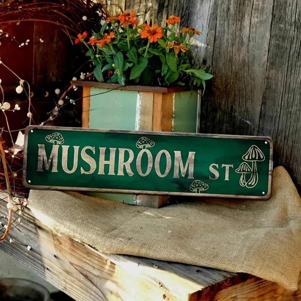 A vintage green metal tin sign with the text Mushroom St is displayed on a wooden surface next to potted flowers.