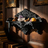 An ornate black Gothic wall shelf displaying three polished mineral specimens on a dark wooden panel.