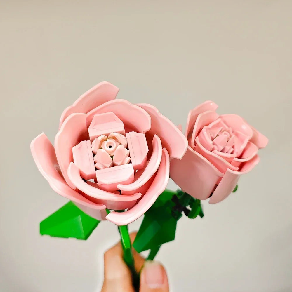 A hand holds a pink DIY rose flower building block bouquet with green leaves against a light background.