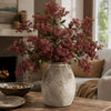 A faux branch of artificial nandina berry stem with dusty plum colored berries in a textured vase on a wooden table.