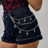 A person wearing dark denim shorts with a layered silver charm belt featuring star and cross pendants.
