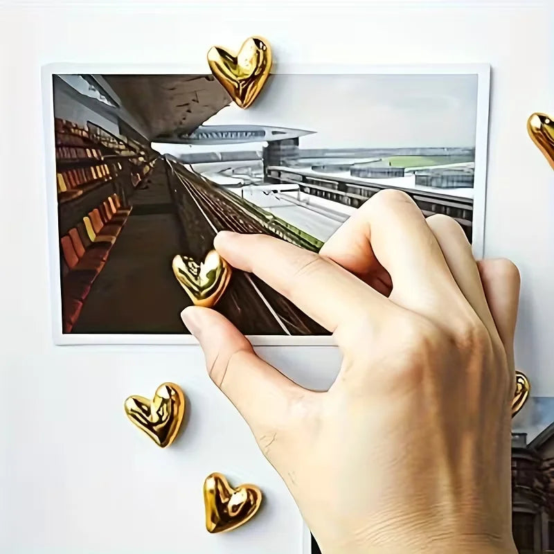 Eine Hand berührt ein gerahmtes Foto eines architektonischen Innenraums mit goldfarbenen herzförmigen Kühlschrankmagneten