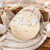 A round wooden birth announcement sign with the word Hello and decorative elements resting on a woven basket and knitted fabr