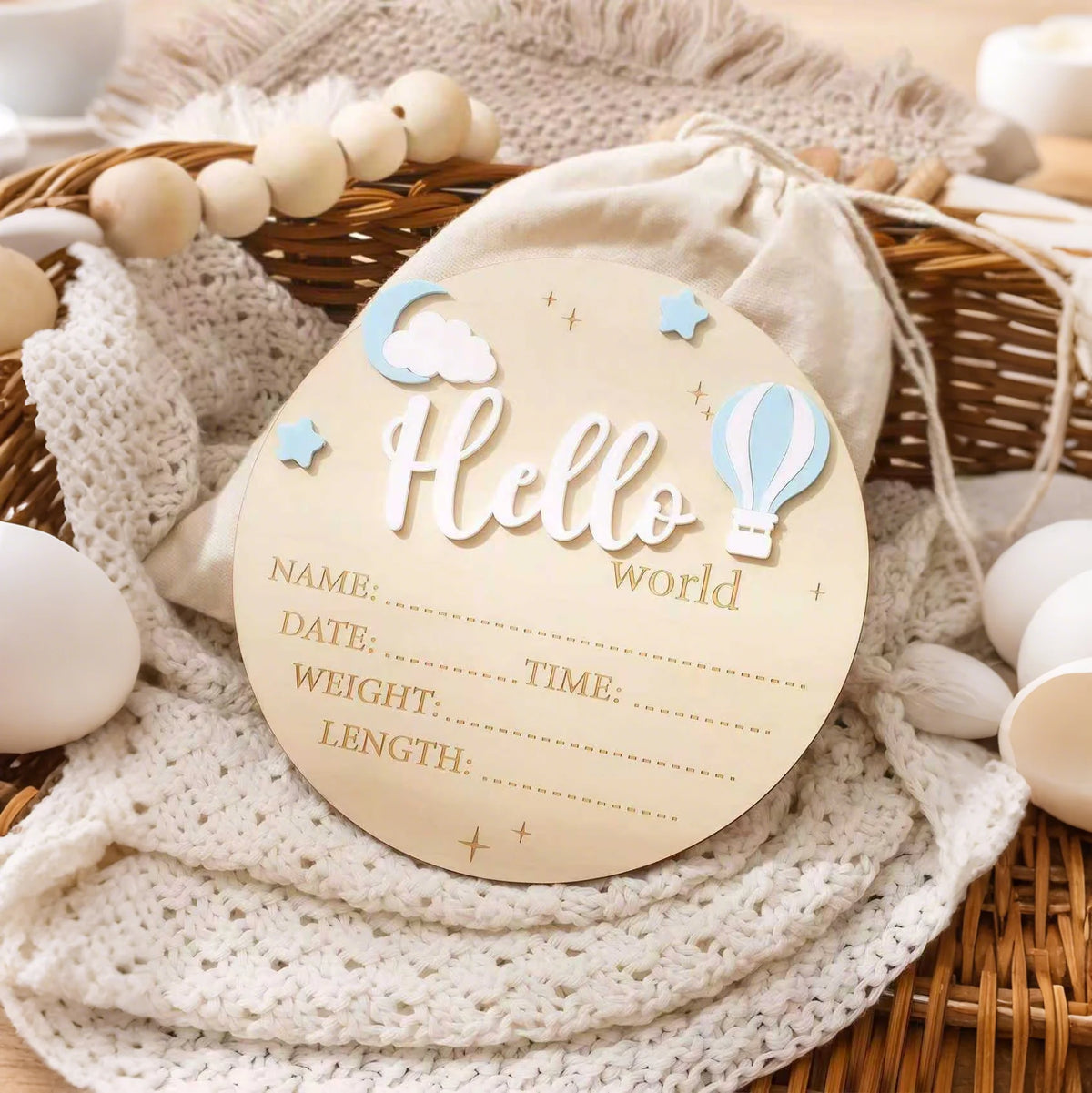 A round wooden birth announcement sign with the word Hello and decorative elements resting on a woven basket and knitted fabr