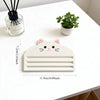 A white cat-shaped squeezer tool for dispensing cat food, placed on a white table next to a potted plant and a glass containe