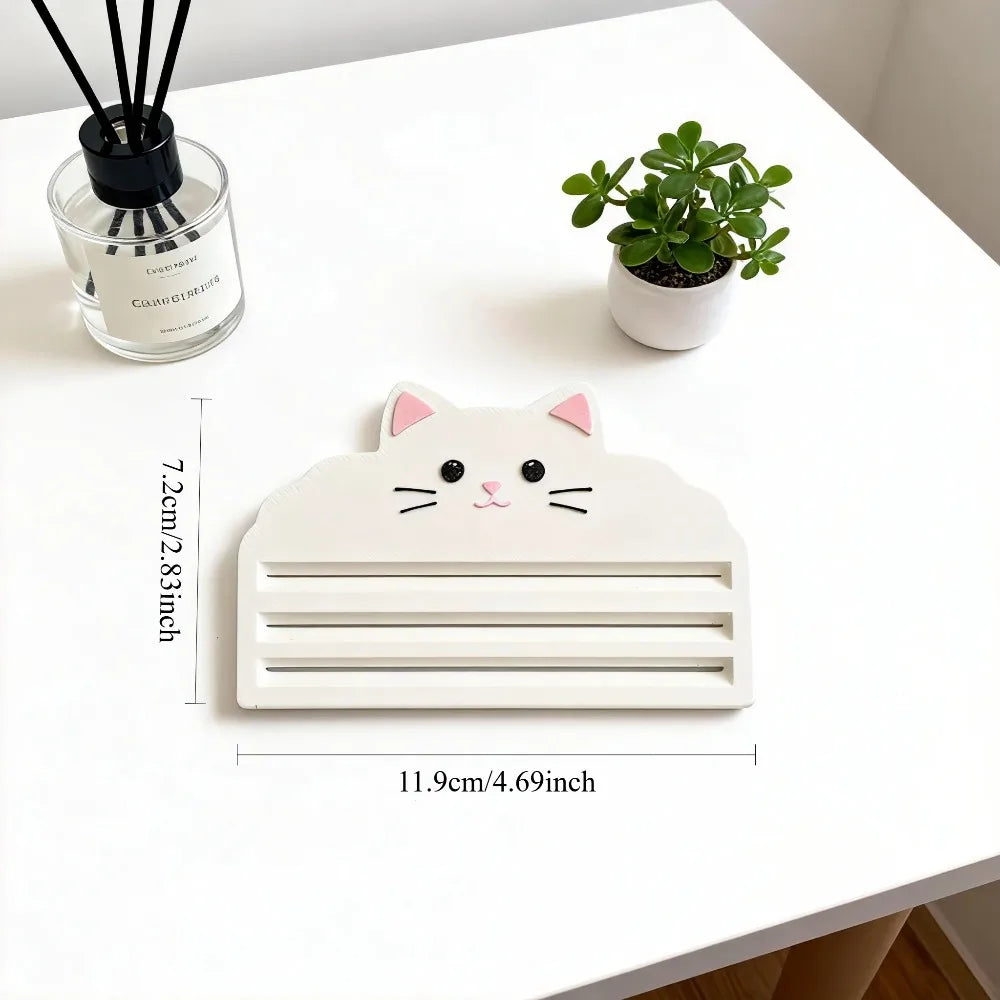 A white cat-shaped squeezer tool for dispensing cat food, placed on a white table next to a potted plant and a glass containe