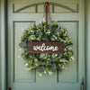 An artificial eucalyptus wreath with a wooden welcome sign hanging on a light green door.