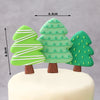 Three green Christmas tree shaped fondant decorations with white stripes and leaves on a white cake.