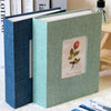 A green linen cover photo album with a floral design and a blue textured cover standing on a white surface.