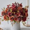 A vibrant bouquet of artificial silk flowers in shades of deep red, burnt orange, and burgundy arranged in a white vase.