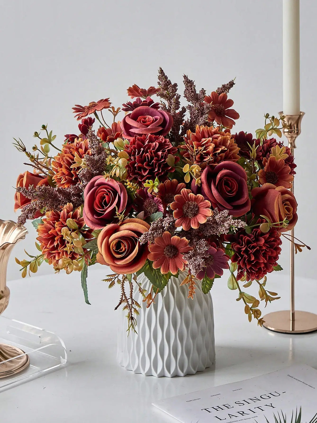 A vibrant bouquet of artificial silk flowers in shades of deep red, burnt orange, and burgundy arranged in a white vase.