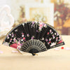 A black lace hand fan with pink and purple floral embroidery resting on a light wooden surface.