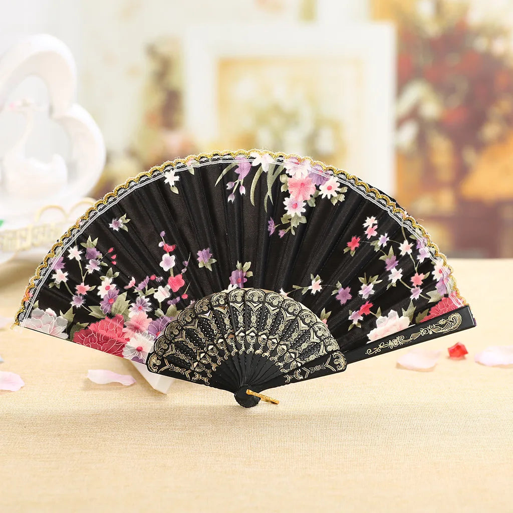 A black lace hand fan with pink and purple floral embroidery resting on a light wooden surface.
