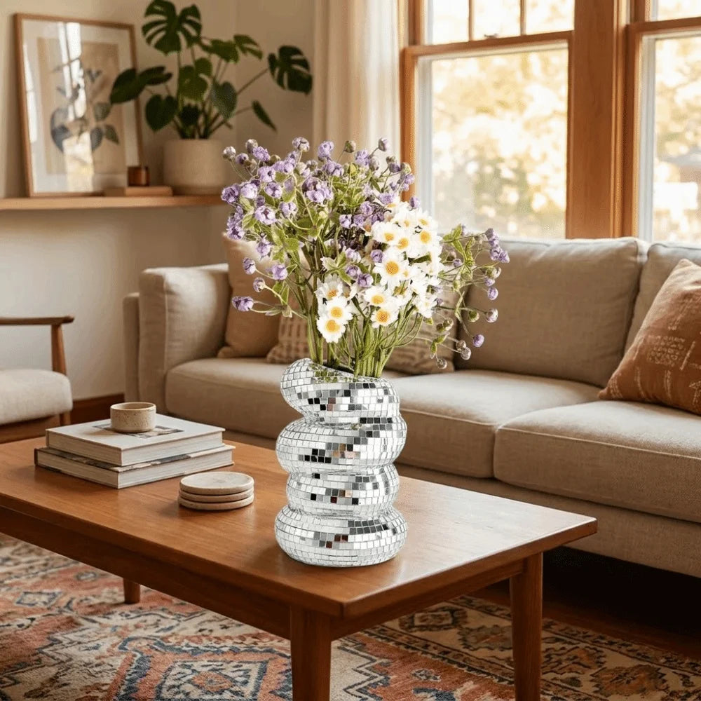 A silver disco ball spiral vase filled with purple and white flowers sits on a wooden coffee table.