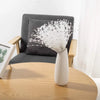 A white ceramic vase filled with delicate pearl string beads sits on a light wooden table next to a gray armchair.