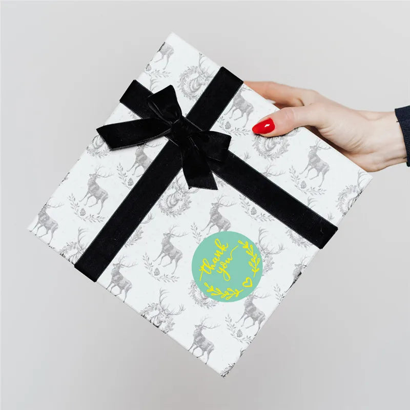 A hand is holding a white gift box tied with a black ribbon, featuring deer silhouettes and a thank you heart sticker.