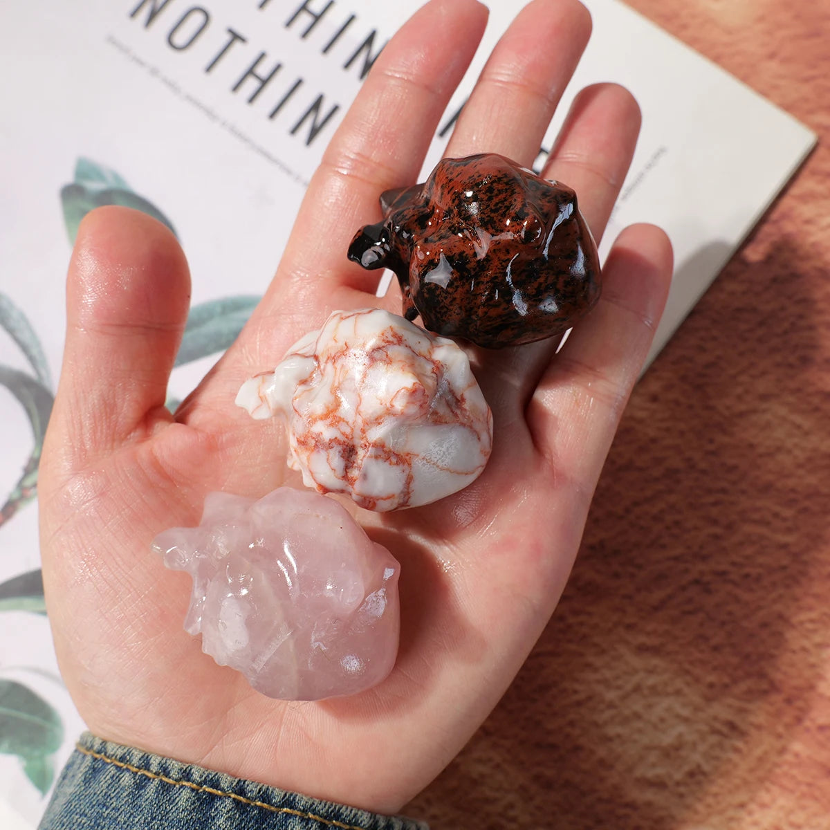 A hand holds three natural crystal heart carvings: a dark reddish-brown, a white and reddish-brown, and a clear pinkish-white