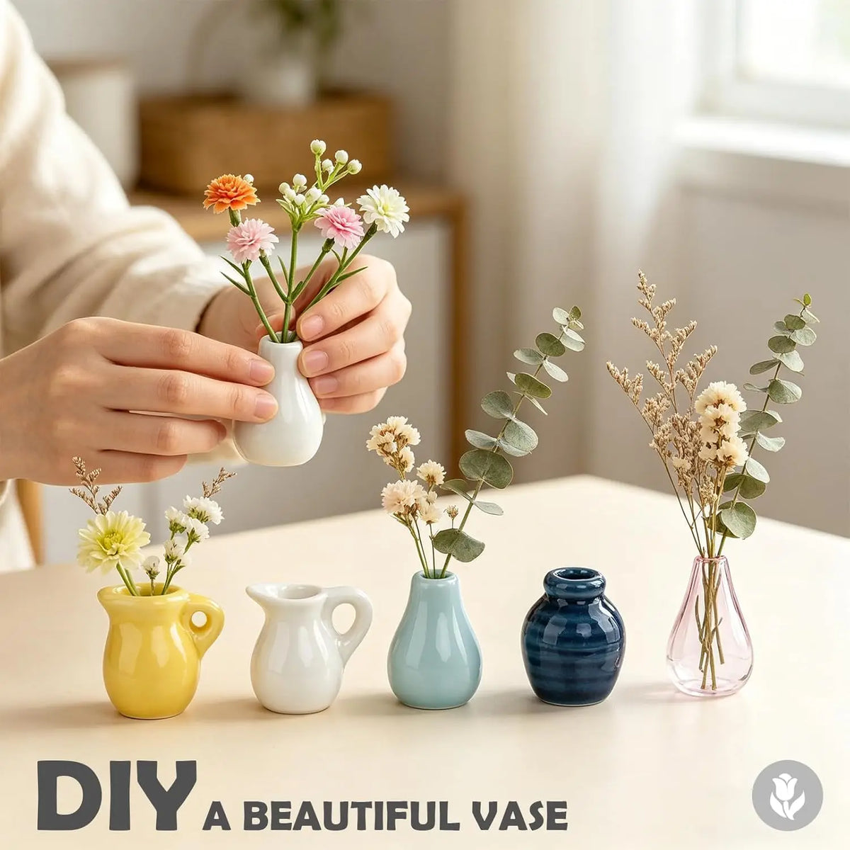 A hand holds a white ceramic vase with pink flowers next to various small blue and yellow ceramic vessels.