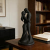 A dark bronze sculpture of a romantic couple embracing and kissing, displayed on a wooden table.
