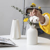A hand is holding a yellow flower in a white ceramic vase on a table.