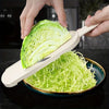 A hand is using a manual cabbage shredder with stainless steel blades to shred a head of green cabbage onto a plate.