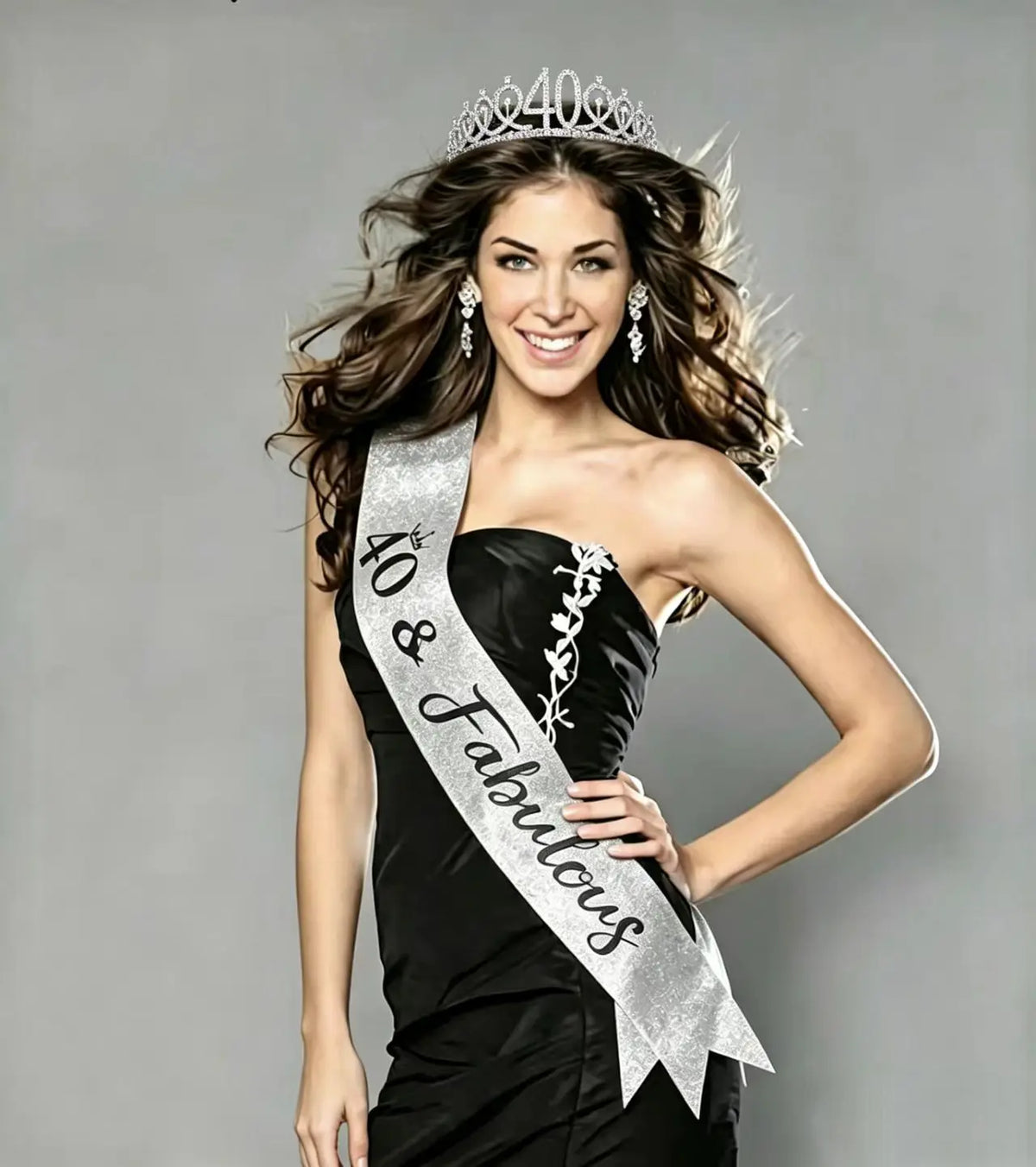 A smiling woman wearing a silver rhinestone birthday crown and glitter sash against a gray background.