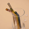 A clear glass contains reusable borosilicate straws and a cleaning brush against a beige background.