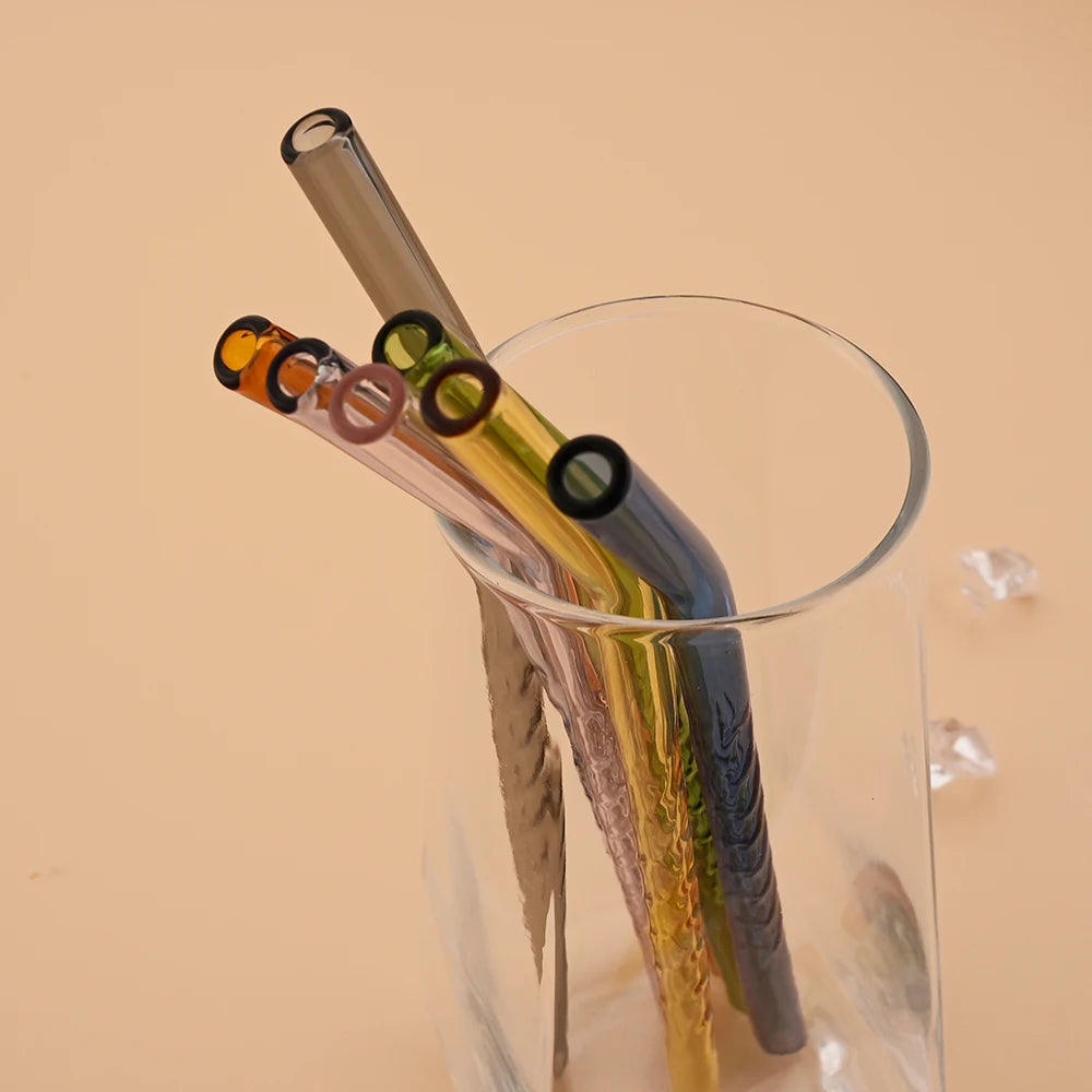 A clear glass contains reusable borosilicate straws and a cleaning brush against a beige background.