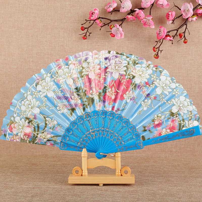 A light blue silk flower folding fan decorated with pink and white floral patterns, displayed on a wooden stand.
