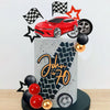 A cake topper set featuring a red racing car, checkered flags, and wheels on a gray cake display.