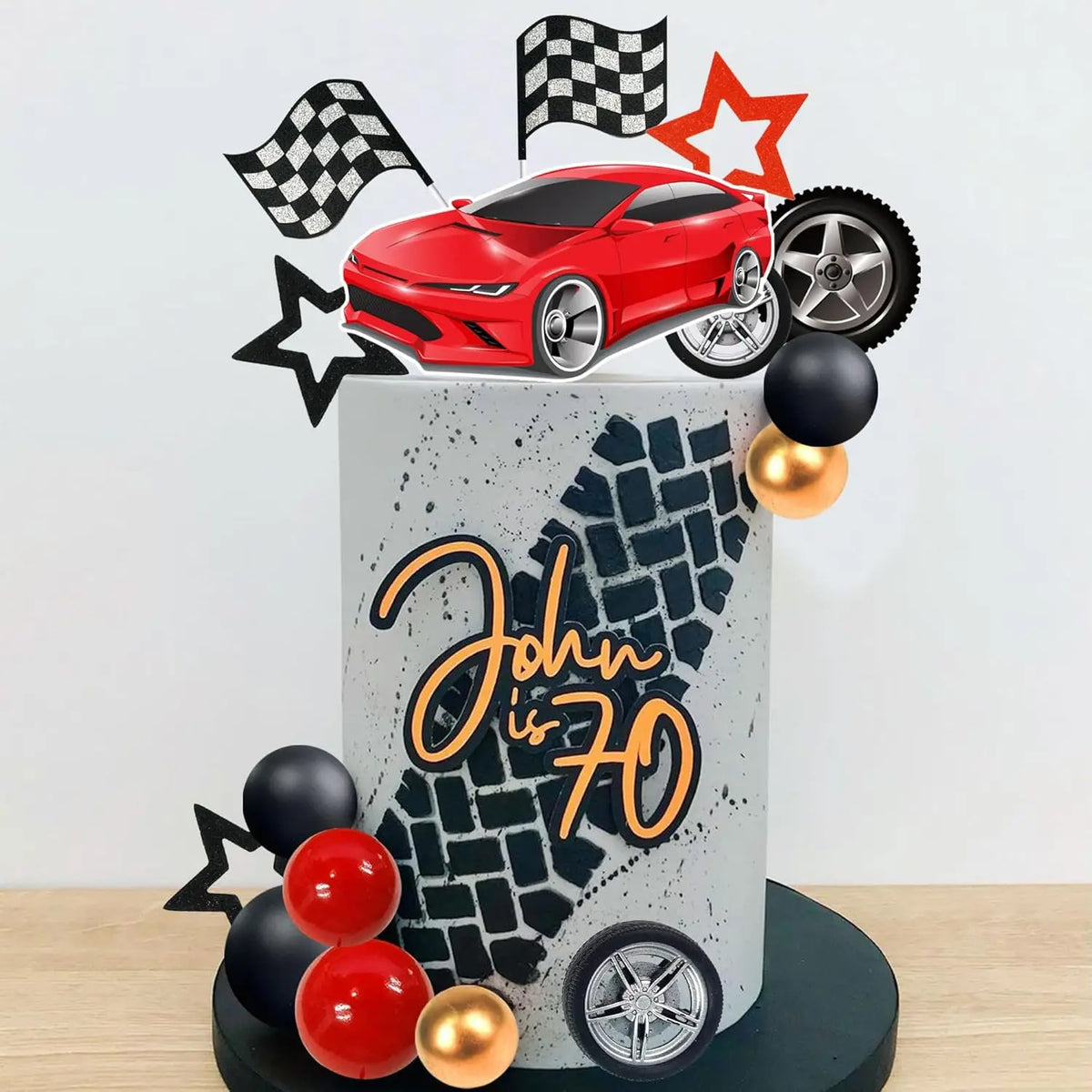 A cake topper set featuring a red racing car, checkered flags, and wheels on a gray cake display.